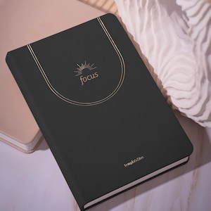May include: A black notebook with the word "focus" in gold script, encircled by a gold line design. The notebook has a gold spine and the word "templatables" in gold at the bottom. A light pink notebook is partially visible in the background.