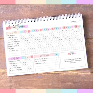 Może przedstawiać: Spiralnie oprawiony notatnik do śledzenia nawyk&oacute;w o kolorowym designie. Okładka zawiera napis "Habit Tracker" w ozdobnej czcionce. Wewnątrz znajduje się układ siatki do śledzenia codziennych, tygodniowych i miesięcznych nawyk&oacute;w.