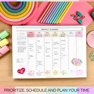 May include: A weekly planner with a pink background, featuring a detailed schedule layout. The planner includes sections for each day of the week, with space for notes and appointments. Colourful pencils and highlighters are arranged around the planner.