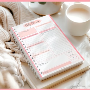 May include: A spiral-bound daily planner with a pink and white design. The planner features sections for priorities, to-do lists, and a brain dump area. The words "Daily Planner" are written at the top. A mug of liquid and a sweater are in the background.