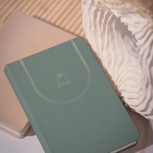 May include: A sage green notebook with the word "focus" in gold script, framed by a gold arc design. A second, light pink notebook is partially visible in the background. The notebooks are likely for journaling or planning.