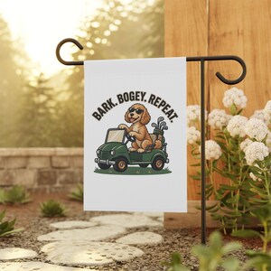 May include: A white garden flag featuring a cartoon illustration of a brown dog wearing sunglasses, driving a green golf cart with a golf bag in the back. The text above the dog reads BARK. BOGEY. REPEAT. in bold black capital letters.