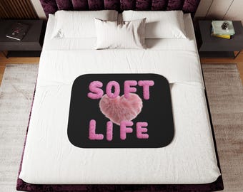 Può includere: Una coperta nera con le parole "SOFT LIFE" in lettere rosa e soffici e un cuore rosa al centro. La coperta è su un letto bianco con una testiera viola e due comodini neri.