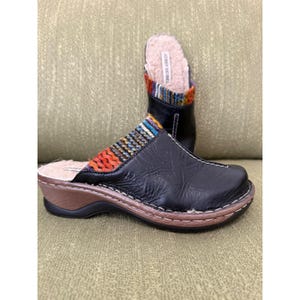 Pode incluir: Tamancos de couro preto com cabedal têxtil tecido colorido e forro de pele sintética. Os tamancos têm sola de madeira e são projetados para conforto e estilo. A marca "JOSEF SEIBEL" é visível.