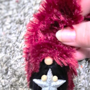 Pode incluir: Enfeite de gnomo feito à mão com chapéu de pele sintética bordô e barba preta. O rosto do gnomo apresenta uma estrela de glitter prateado e duas pequenas esferas bege. Enfeite para decoração de feriados ou sazonal.