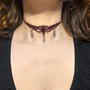 Könnte beinhalten: Ein burgunderfarbenes Makramee-Choker-Halsband mit goldfarbenen Perlen und drei herabhängenden tropfenförmigen Anhängern. Die Halskette hat einen zentralen lila Edelstein und wird um den Hals getragen.