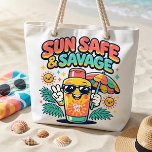 May include: White canvas tote bag with rope handles. The bag features a colorful graphic of a sunscreen bottle wearing sunglasses and holding a beach umbrella. Text on the bag reads "SUN SAFE & SAVAGE". The bag is on a sandy beach.