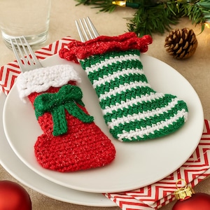 May include: Two crocheted Christmas stocking fork holders. One is red with a white cuff and green bow. The other is green and white striped with a red cuff. They sit on white plates with red and white patterned napkins.
