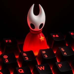 May include: A figurine of a character with a white head and a red cape, adorned with a gold star, stands on a black keyboard. The keys are black with red illuminated characters. The background is dark.