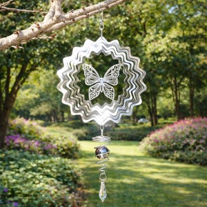 May include: A silver metal wind spinner with a butterfly design suspended from a tree branch. The spinner features a layered, wavy outer ring and a central butterfly cutout. A spiral and crystal pendant hang below.
