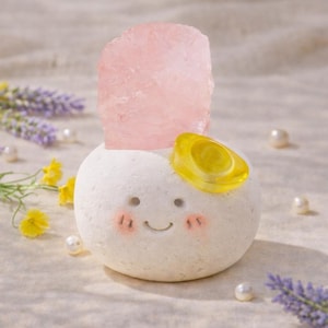 May include: A charming, white, spherical sculpture with a smiling face, pink cheeks, and a yellow detail. A large, roughly-cut, pink crystal is placed on top. Pearls and lavender sprigs are scattered around the sculpture.