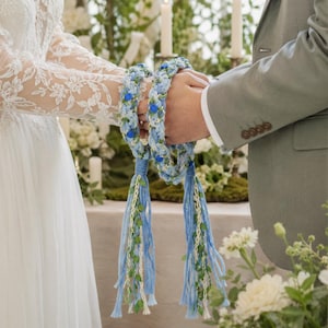 Puede incluir: Un cordón de handfasting azul claro y blanco con detalles florales azules y largas borlas. El cordón es sostenido por dos personas, una con un vestido de encaje blanco y la otra con un traje gris. Un símbolo de unidad en una ceremonia de boda.