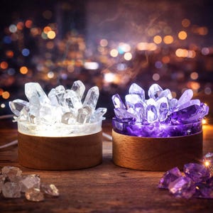 May include: Two crystal cluster lamps with wooden bases. One lamp features clear crystals, the other purple crystals. The lamps are illuminated, creating a soft glow. Crystals are scattered on the wooden surface.