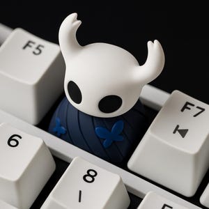 May include: A white and blue custom keycap shaped like a character from a video game, with a white head and black eyes. The keycap is on a white keyboard, with the F5, 6, 8, and F7 keys visible.