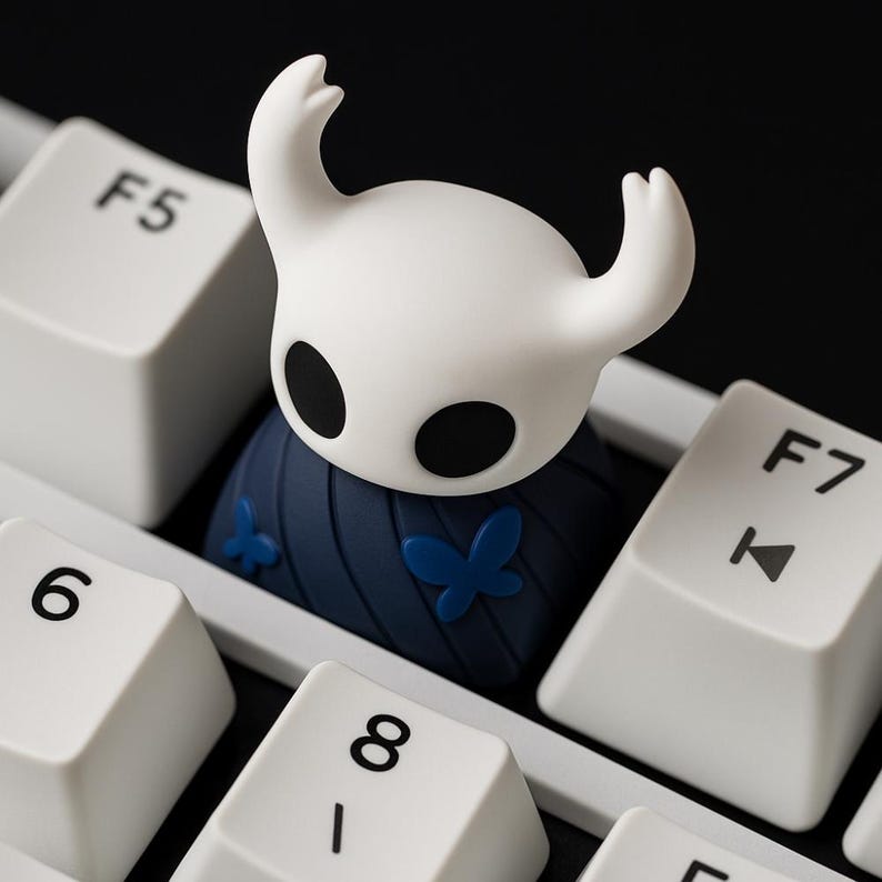 May include: A white and blue custom keycap shaped like a character from a video game, with a white head and black eyes. The keycap is on a white keyboard, with the F5, 6, 8, and F7 keys visible.