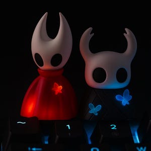 May include: Two stylized figures with glowing butterfly accents. One figure wears a red cape, the other a black cloak. Both have white heads with large eye-like openings. The figures are illuminated with red and blue light.