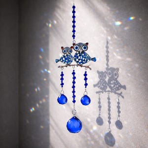 May include: A blue and silver owl suncatcher with two owls perched on a branch. The owls have blue and white patterned wings and large blue eyes. Below the owls are three blue crystal balls and a large faceted crystal. The suncatcher is hanging.