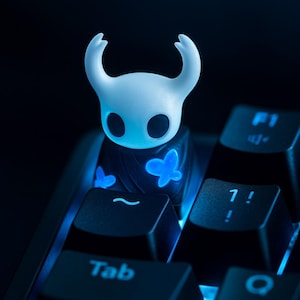 May include: A white, horned character keycap with glowing blue butterfly accents, resting on a black keyboard. The keycap has a unique design and is illuminated with blue light, creating a striking visual effect. The keyboard keys are also backlit.