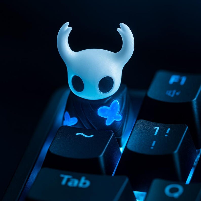 May include: A white, horned character keycap with glowing blue butterfly accents, resting on a black keyboard. The keycap has a unique design and is illuminated with blue light, creating a striking visual effect. The keyboard keys are also backlit.