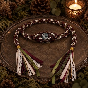 May include: A braided cord belt featuring a central oval moss agate stone, crafted from intertwined burgundy, olive green, and white threads. The ends are finished with decorative tassels made of matching ribbons, lace, and metallic gold trim.