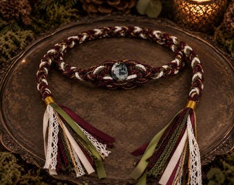 May include: A braided cord belt featuring a central oval moss agate stone, crafted from intertwined burgundy, olive green, and white threads. The ends are finished with decorative tassels made of matching ribbons, lace, and metallic gold trim.