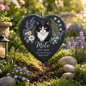 Puede incluir: Una placa conmemorativa de pizarra negra en forma de corazón con el retrato de un gato blanco y negro rodeado de ilustraciones florales. El texto dice Milo, 2017-2025, y Forever In Our Hearts, con una estaca de metal para el jardín.