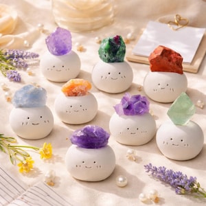 May include: A selection of white ceramic ornaments, each adorned with a vibrant gemstone. The gemstones include amethyst, emerald, orange calcite, and red jasper. The ornaments feature smiling faces and are displayed on a light-toned surface.