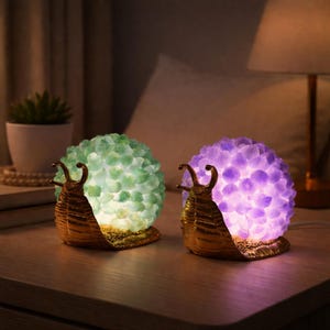 May include: Two snail-shaped lamps with illuminated, textured bodies. One lamp has a green body, the other is purple. The snail bodies are made of a crystal-like material, and the base is gold-coloured.