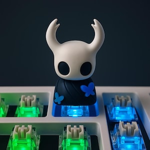 May include: A white and black figurine with curved horns and large black eyes, adorned with blue butterfly accents, sits atop a keyboard key. The key is illuminated with blue light, and other keys glow green. The figurine has a black cape.