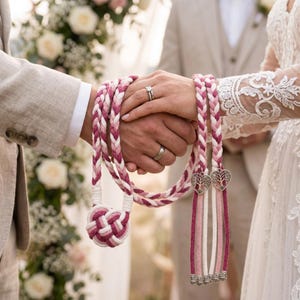 Pode incluir: Um cordão de união de mãos com cordas trançadas em tons de branco, rosa claro e rosa escuro. O cordão inclui um nó decorativo de punho de macaco numa extremidade e dois berloques prateados em forma de coração com borlas de corda pendentes.