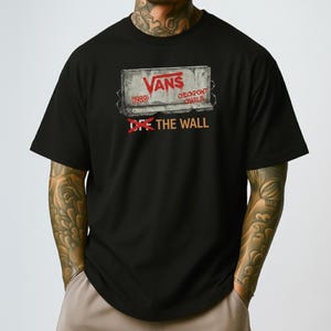 Könnte beinhalten: Schwarzes T-Shirt mit einem grafischen Design mit dem Vans-Logo und dem Text "Checkpoint Charlie (1989)". Der Schriftzug "Off The Wall" ist in Orange auf dem T-Shirt aufgedruckt.