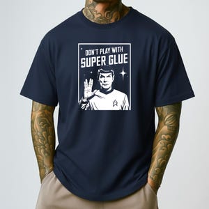 Puede incluir: Camiseta azul marino con un gráfico blanco que presenta un personaje de Star Trek y el texto "DON'T PLAY WITH SUPER GLUE". El gráfico incluye un gesto con la mano y una insignia de Starfleet.