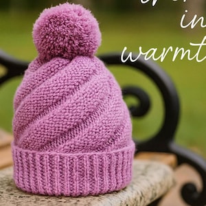 May include: A lilac knitted hat with a pom-pom rests on a bench. The hat has a ribbed band and a diagonal cable knit design. The words "Wrap them in warmth" are written in white script. The website www.crochix.com is also visible.