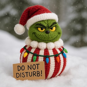 May include: A crocheted Grinch figure wearing a red and white striped sweater, a Santa hat, and a string of colorful lights. The Grinch has a green face with a mischievous grin and yellow eyes. A small sign reads "DO NOT DISTURB!"
