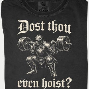 K&ouml;nnte beinhalten: Schwarzes T-Shirt mit einer Grafik eines Ritters in R&uuml;stung, der eine Langhantel hebt. Der Text "Dost thou even hoist?" ist oberhalb und unterhalb der Ritterillustration in gotischer Schrift gedruckt. Das T-Shirt hat ein Comfort Colors-Etikett.