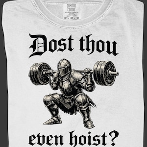 K&ouml;nnte beinhalten: Wei&szlig;es T-Shirt mit einem schwarzen Grafik eines Ritters, der Gewichte hebt, mit dem Text "Dost thou even hoist?" in gotischer Schrift. Das Shirt hat einen Rundhalsausschnitt und ein Etikett "Comfort Colors".