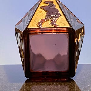 Op de afbeelding: Een decoratieve, facetgeslepen, bordeauxrode glazen doos met een goudkleurig deksel. Het deksel heeft een verhoogd reliëf van een figuur in beweging. De doos heeft een vierkante basis en een geometrisch, schuin ontwerp.