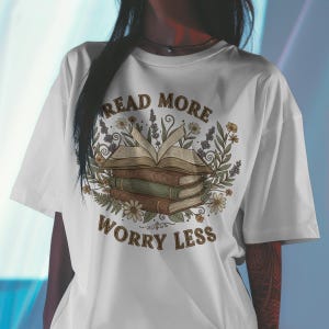 Könnte beinhalten: Weißes T-Shirt mit einem Design, das einen Stapel Bücher, ein offenes Buch und florale Elemente zeigt. Der Text "Read More Worry Less" ist in brauner Schrift über und unter den Büchern angeordnet. Das Design hat eine Vintage- und botanische Ästhetik.