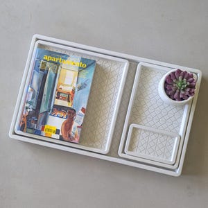 Puede incluir: Un conjunto de cuatro bandejas rectangulares blancas de diferentes tamaños, anidadas. La bandeja más grande contiene una revista con la palabra "apartamento" en la portada. Una pequeña maceta blanca con una suculenta morada se encuentra en una de las bandejas.