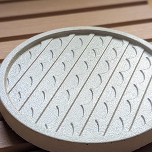 Puede incluir: Una bandeja redonda de hormigón blanquecino con una superficie texturizada. La bandeja presenta ranuras diagonales con recortes semicirculares. La bandeja está sobre una superficie de madera.