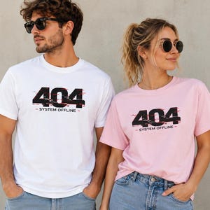 Könnte beinhalten: Ein weißes T-Shirt und ein hellrosa T-Shirt, beide mit einer schwarzen Grafik mit dem Text 404 SYSTEM OFFLINE im Glitch-Stil. Das Design enthält horizontale rote und schwarze Linien über den Zahlen und ist mittig auf der Brust aufgedruckt.