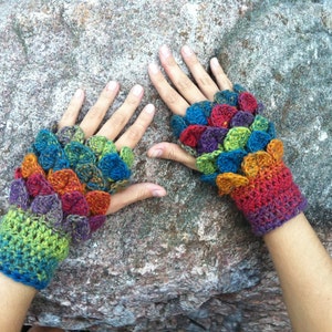 May include: A pair of colourful crocheted fingerless gloves with a scale pattern. The gloves are made with a variety of colours, including red, orange, yellow, green, blue, and purple.