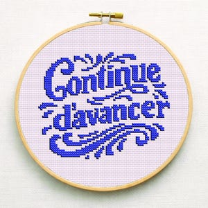 May include: A cross-stitch embroidery hoop with the words "Continue d'avancer" in blue thread on a light purple fabric. The text is surrounded by decorative flourishes. The wooden hoop has a brass adjustment screw.