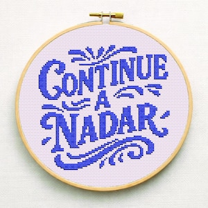 May include: A cross-stitch embroidery hoop featuring the words "CONTINUE A NADAR" in blue on a light purple background. The text is in a decorative font. The wooden hoop has a brass clasp.