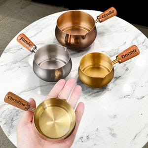 May include: Four small, metal saucepans with wooden handles. The pans are in copper, silver, and gold tones. One pan is held in a hand. The wooden handles are engraved with names: Jamie, Giselle, Donna, and Christine.