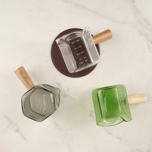 May include: Three glass measuring cups with wooden handles. One clear, one gray, and one green. The clear cup is on a brown coaster. The clear cup has measurements in ounces and milliliters. The gray cup has the name "Graham" etched on the handle.