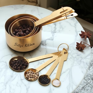 May include: A set of gold-colored measuring cups and spoons. The cups are nested, with coffee beans inside. The spoons are filled with peppercorns, salt, and other spices. The text "Betsy's Kitchen" is engraved on the largest cup.
