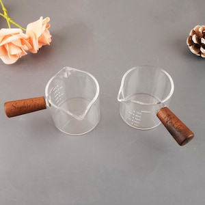 May include: Two clear glass measuring cups with wooden handles. One handle is engraved with "Sophie's", the other with "Nahla". Each cup has measurement markings and a pouring spout. The cups are on a gray surface.