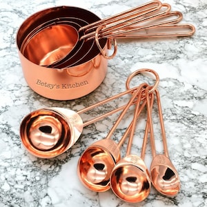 May include: A set of copper measuring cups and spoons. The measuring cups are nested together, with the text "Betsy's Kitchen" engraved on the side. The spoons are on a ring. The set is made of copper and sits on a marble surface.