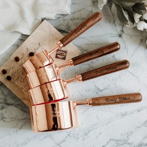 May include: A set of four copper measuring cups with wooden handles. Each cup is engraved with a name or phrase: Lesley, Mindtrang, Carl's Kitchen, and Homely Kitchen. The cups are stacked, showing measurements.